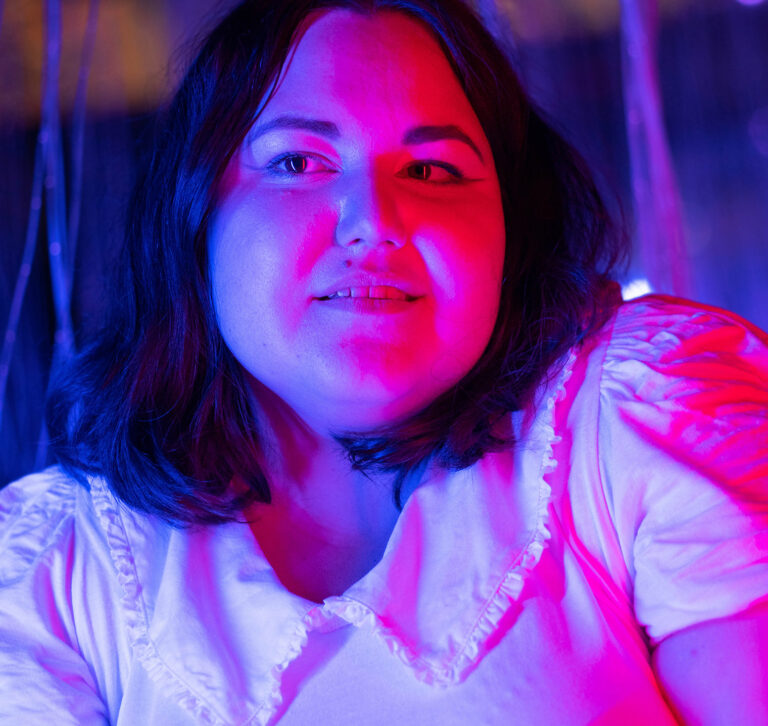Portrait einer braunhaarige Frau in weißem T-Shirt mit Kragen wird rechts von rotem und links von blauem Licht angestrahlt.