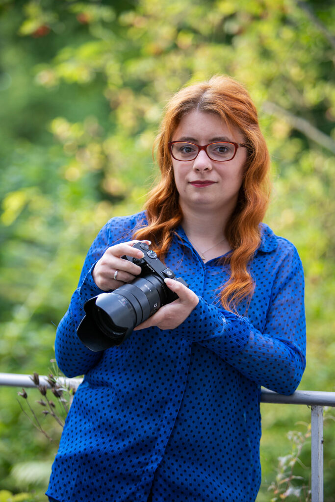 Die Inhaberin der Webseite Katharina Hofer steht mit naturroten Haaren, einer blauen Bluse mit dunkelblauen Punkten mit ihrer Kamera in der Hand lächelnd an ein Geländer gelehnt.