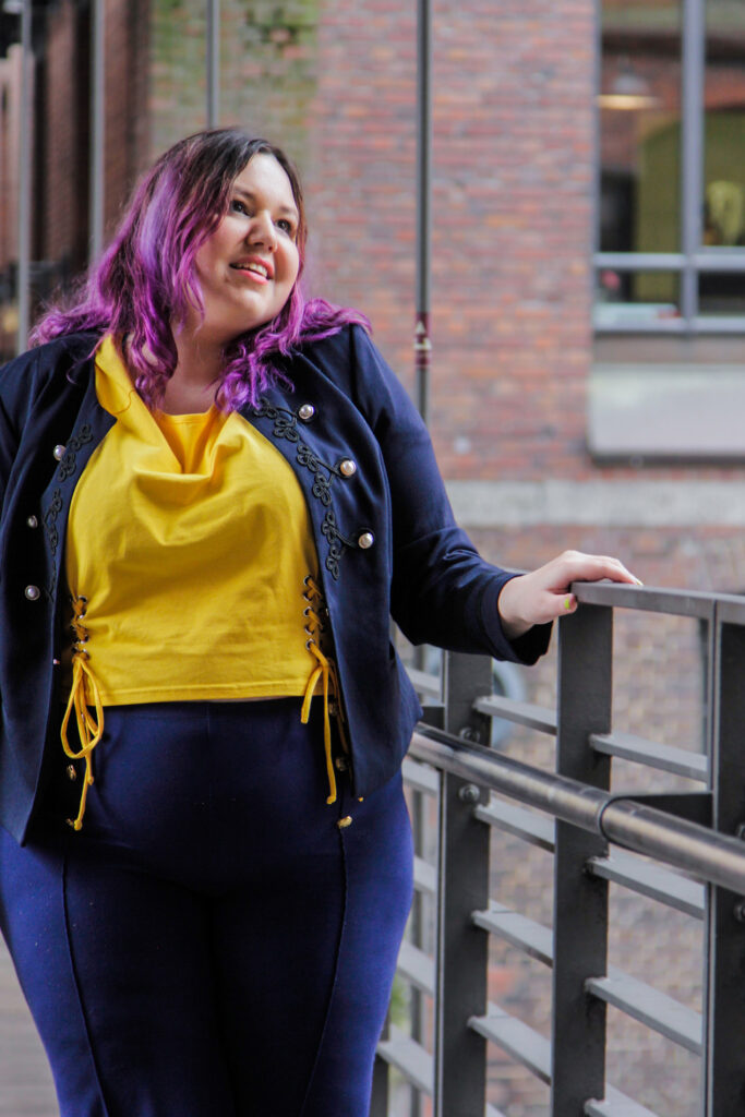 Eine Frau mit braunen Haaren und lila Spitzen steht in Hamburg in der Speicherstadt unter ein Brücke ans Geländer gelehnt. Sie hat eine Dunkelblaube Jacke mit goldenen Knöpfen, eine passende Hose und ein gelbes T-Shirt an.