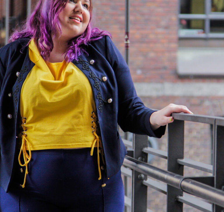 Eine Frau mit braunen Haaren und lila Spitzen steht in Hamburg in der Speicherstadt unter ein Brücke ans Geländer gelehnt. Sie hat eine Dunkelblaube Jacke mit goldenen Knöpfen, eine passende Hose und ein gelbes T-Shirt an.