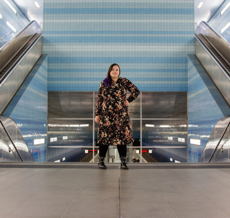 Eine braunhaarige mit lila Spitze Frau steht in einem Blumenkleid in einer Hamburger U-Bahnstation. Diese Hat blau geachtelte Wände und erinnert vom Design an ein Schwimmbad.